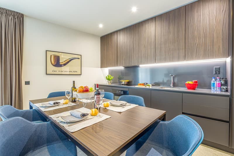 Modern dining room with wooden dining table and blue chairs, wooden cabinets, counter with sink and kitchen appliances, bowl of fruits, wine bottles, wine glasses, and other dining items