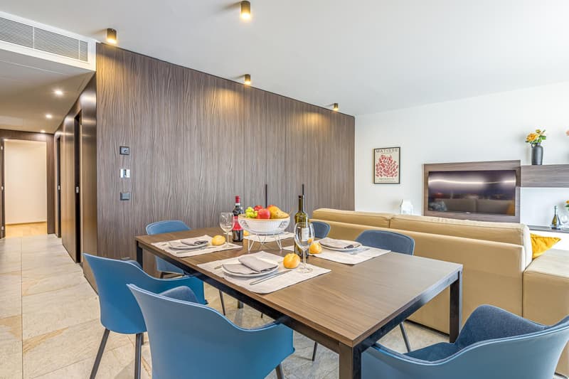 Dining table, wine bottles, fruit bowl, blue chairs, beige sofa, television, flowers