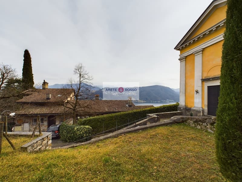 A yellow-colored building with a classical architectural style, surrounded by a grassy area and trees. The building has a large sign that says 'ABETE ROSSO' on it. There are other smaller buildings and a car visible in the background, with mountains in th