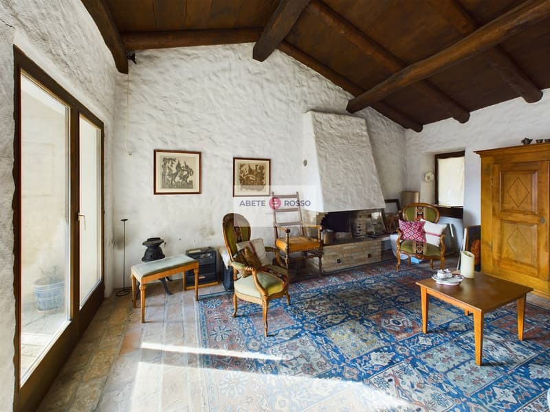 Rustic living room with exposed wooden beams, white stone walls, antique furniture including armchairs and a coffee table, and a large patterned area rug. There is a fireplace visible in the room.
