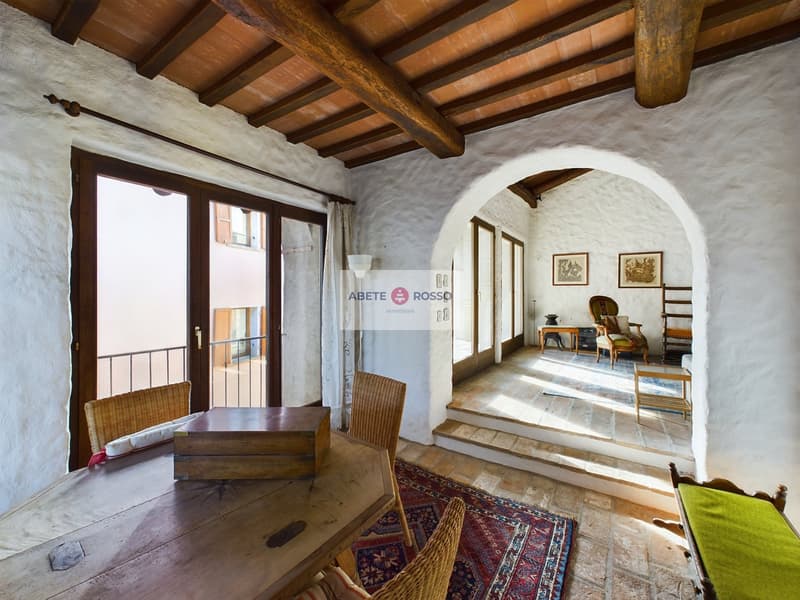 Rustic living room with wooden beamed ceiling, arched doorway, wooden furniture, and a balcony or terrace visible through the window.