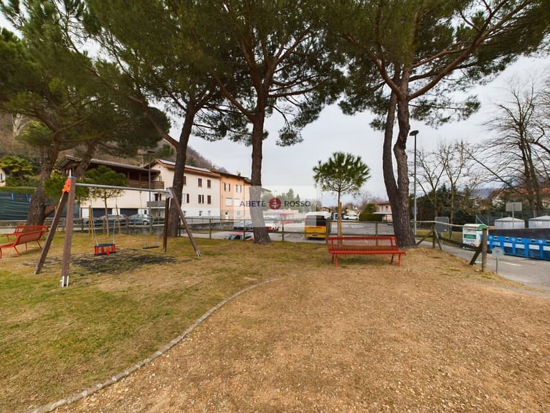 Outdoor park area with playground equipment, benches, and trees. There are apartment buildings visible in the background.
