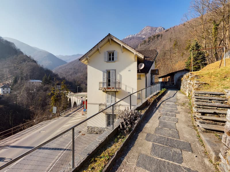 Two-story house with yellow exterior, tiled roof, balcony, surrounded by mountains and trees, with a stone pathway leading up to the house