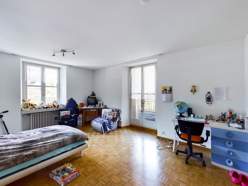 Spacious bedroom with large windows, parquet flooring, a bed, a sofa, a desk with a chair, and various personal items and decorations such as toys, a globe, and wall art.