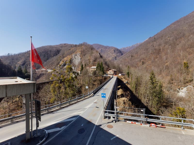 The image shows a scenic mountain landscape with a winding road or bridge crossing a valley. The road is surrounded by forested hills and mountains, with a red flag visible on a structure in the foreground. The landscape appears to be in a mountainous, ru