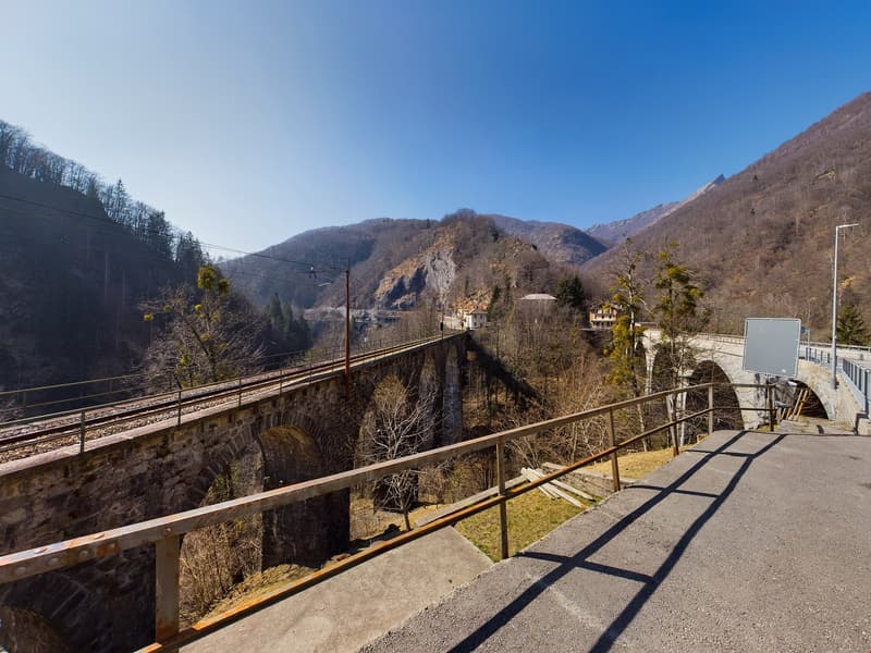 Stone bridge with metal handrails, concrete walkway, surrounded by mountains, power lines, a few trees, streetlight, and a sign