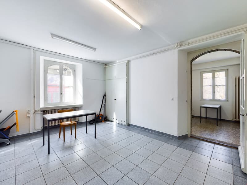 This image shows a large, open room with white walls and a tiled floor. The room has a desk and chairs, as well as a window that provides natural light. The room appears to be a workspace or office space.