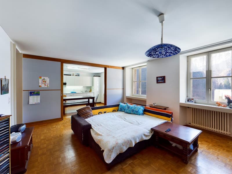 Spacious bedroom with hardwood flooring, large windows, a couch, a wooden coffee table, and a blue and white patterned ceiling light fixture.