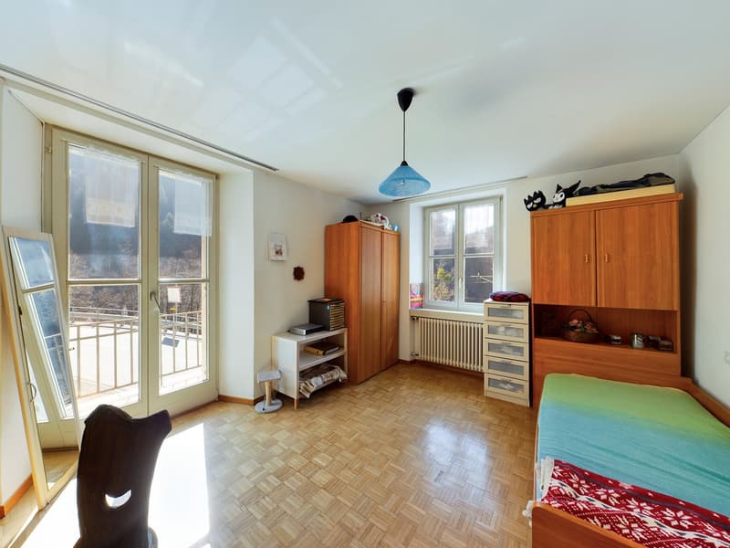 Spacious bedroom with large windows, wooden furniture including a wardrobe and shelving unit, and a balcony or terrace accessible through the windows.