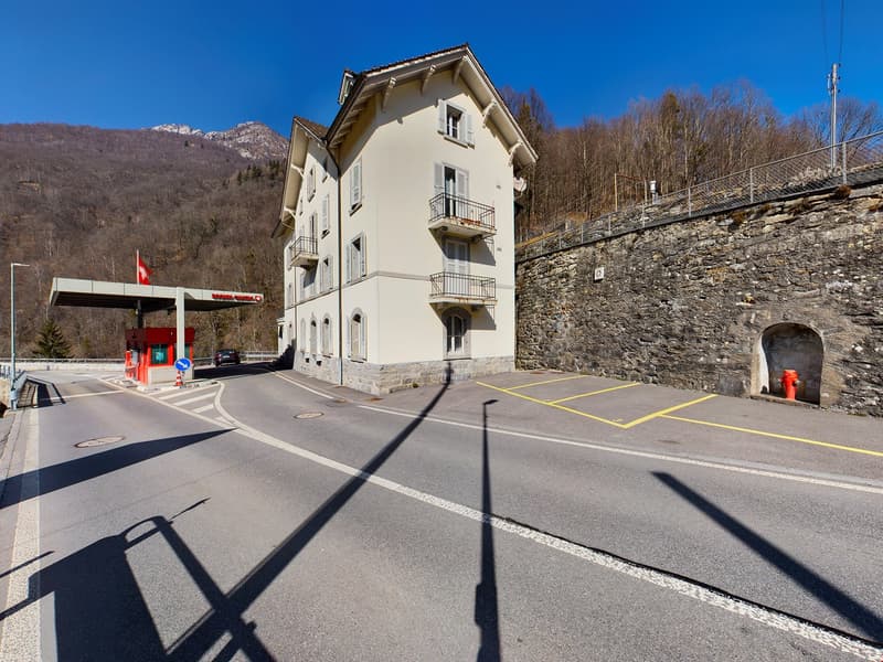 A multi-story building with a stone facade, located in a mountainous area with a clear blue sky. The building has balconies and windows, and there is a road and parking area in front of it.