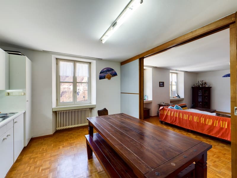 The image shows a room with a large wooden table, a sofa with an orange blanket, and a cabinet in the background. The room has wooden floors and walls, and large windows that let in natural light. The room appears to be a living or dining area.