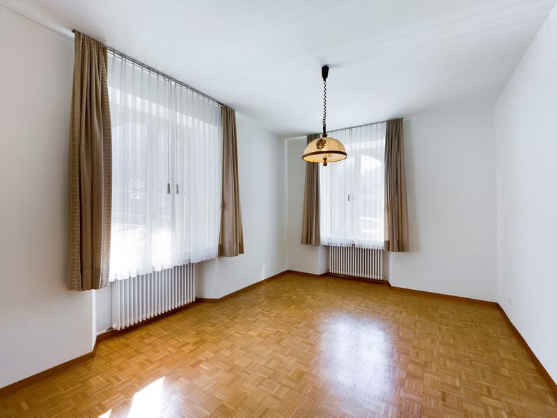 Spacious living room with hardwood flooring, large windows with sheer curtains, and a pendant light fixture. The room appears to be empty, providing a clean and minimalist aesthetic.