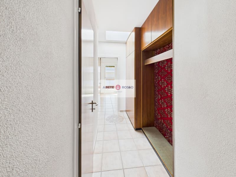 The image shows a hallway with a tiled floor, white walls, and a wooden cabinet or closet along one wall. The cabinet has a red patterned fabric curtain or door covering part of it. The hallway appears to be well-lit and spacious.
