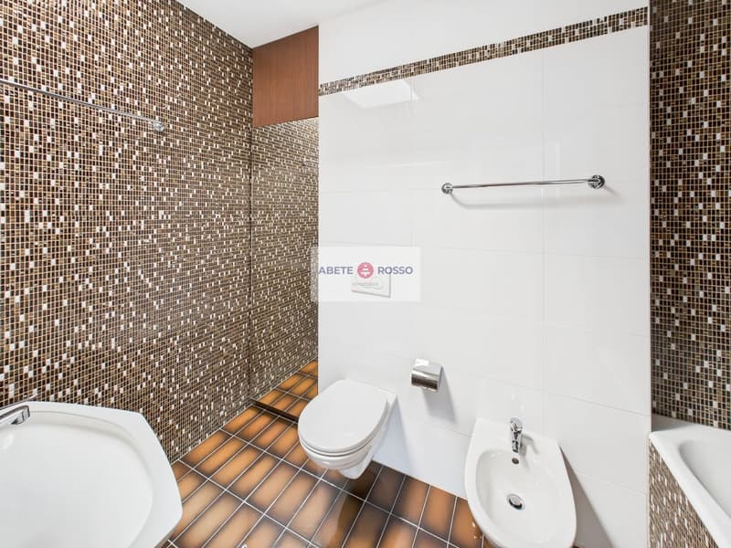 The image shows a modern bathroom with a white toilet, sink, and a tiled wall in a mosaic pattern of brown and beige tiles. There is a towel rack and a Diabete Rosso sign on the wall.