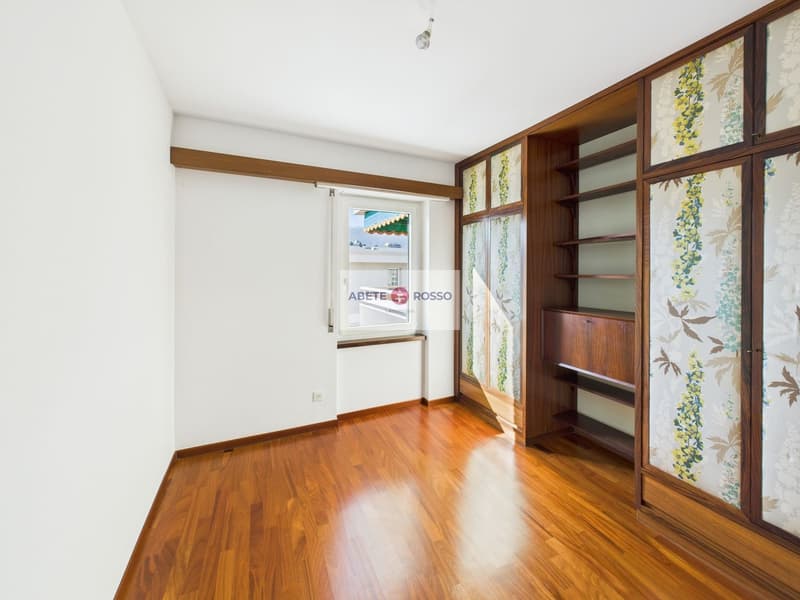 Spacious bedroom with hardwood flooring, wooden shelving units, and floral patterned curtains. There is a window with a view, possibly leading to a balcony or terrace.
