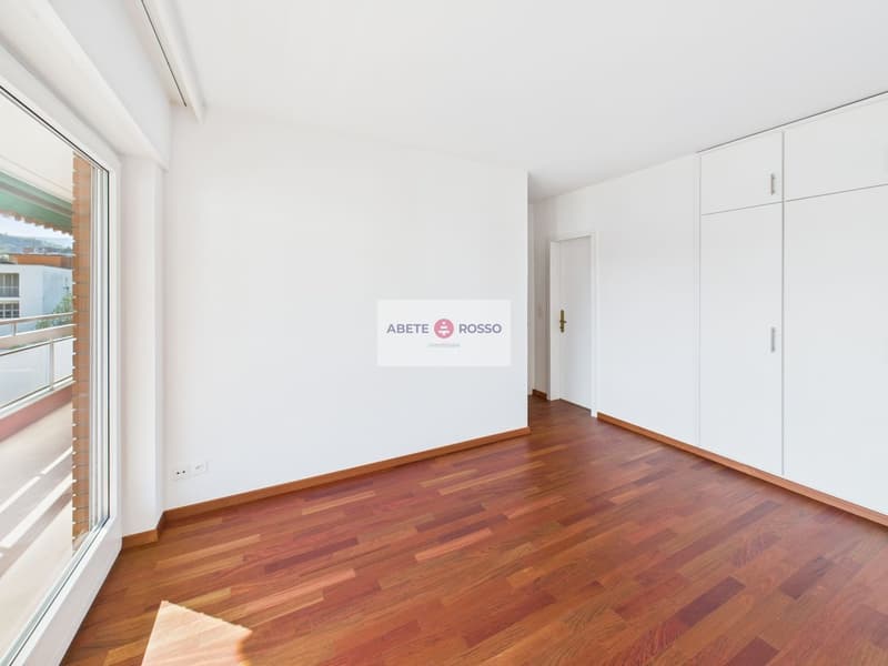 Spacious bedroom with hardwood flooring, large windows providing natural light, and a balcony or terrace visible.