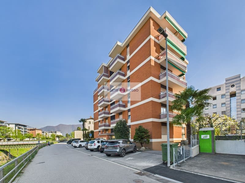 Multi-story brick apartment building with balconies, parking spaces in front, and palm trees in the surrounding area.