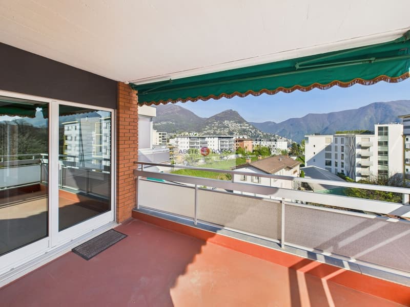Spacious apartment with a large balcony, offering a panoramic view of the surrounding mountains and cityscape. The balcony features a green and gold-colored awning, providing shade and adding to the cozy ambiance. The apartment has large windows, allowing