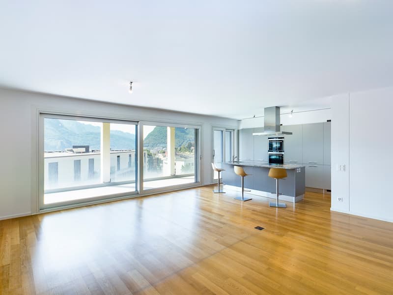 Empty room with wooden floors, large sliding glass windows, kitchen with island, and stools.