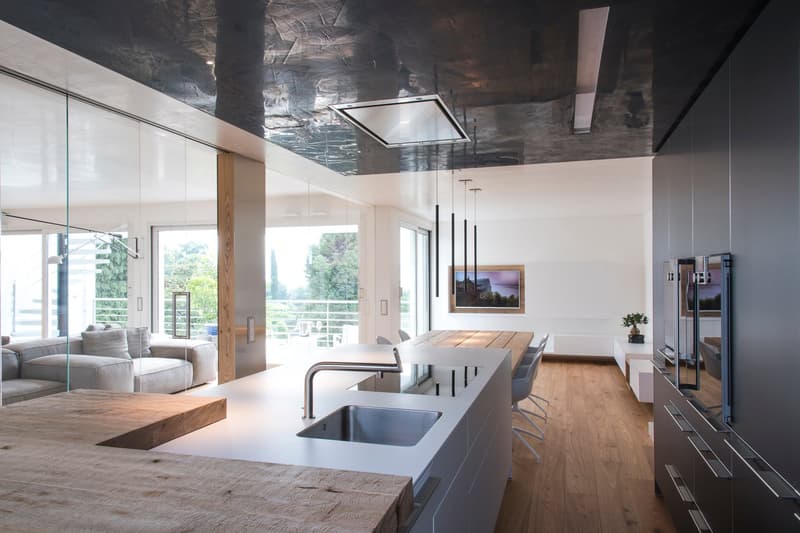 Modern kitchen with wooden countertop, stainless steel sink, faucet, and large windows overlooking a view of a house and trees.