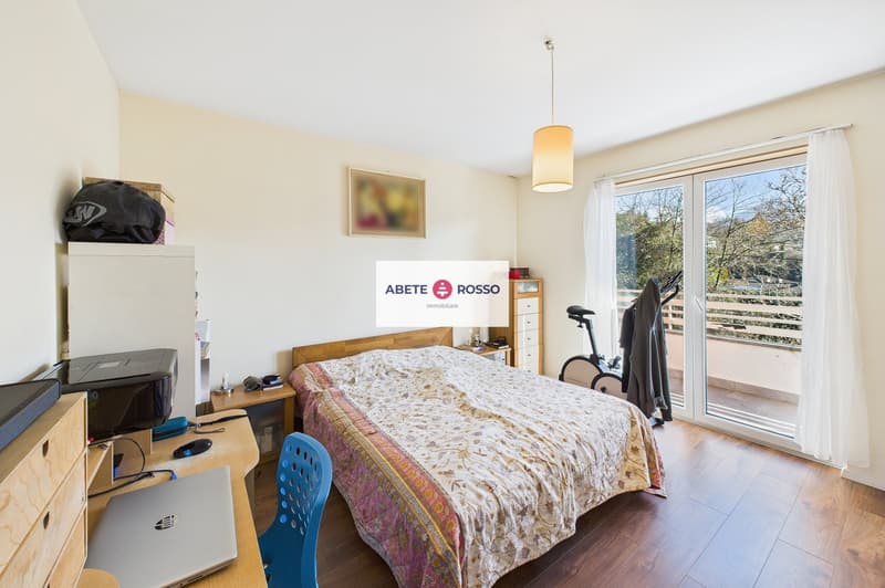 Bedroom with bed, desk, chair, printer, laptop, wooden floor, sliding glass door to balcony, cabinet, picture frame, lamp, exercise bike, clothes hanging
