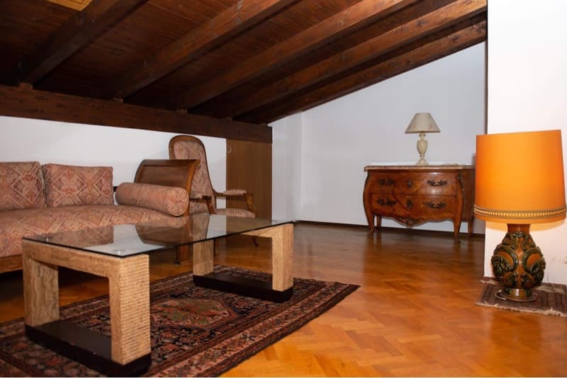 Spacious living room with wooden beamed ceiling, hardwood floors, ornate wooden dresser, glass coffee table, patterned sofa, and a decorative lamp on a side table.