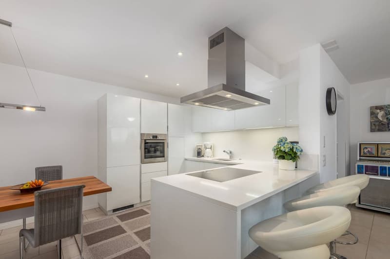 Modern, open-concept kitchen with white cabinets, stainless steel appliances including a range hood, quartz countertops, and a kitchen island with bar stools. The kitchen is connected to a dining area with a wooden table and wicker chairs.