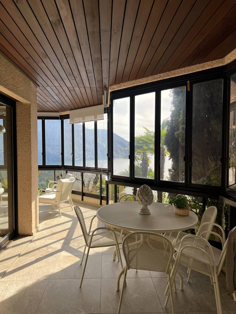 Spacious sunroom with large windows offering a scenic view of the surrounding landscape, including mountains and vegetation. The room features a wooden ceiling, a round dining table with white chairs, and decorative elements like a pineapple ornament.