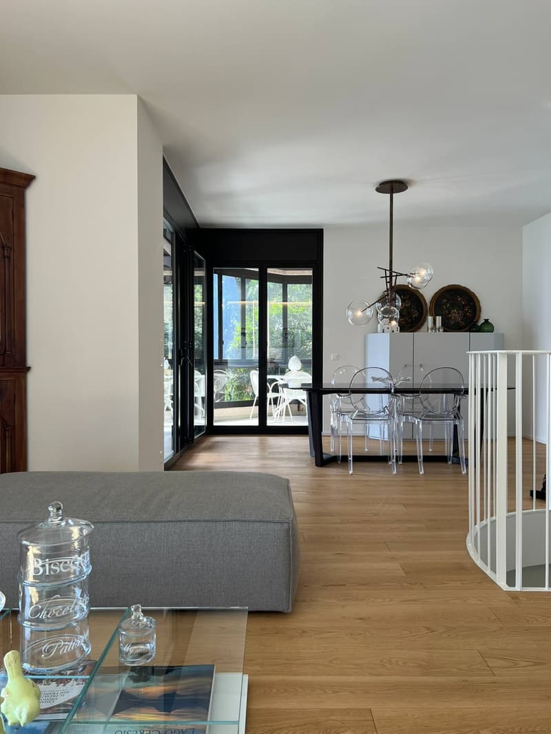 The image shows an open-concept living and dining area with a large glass sliding door leading to a balcony or terrace. The living room features a gray sofa, and the dining area has a glass table with transparent chairs. The room has a modern, minimalist