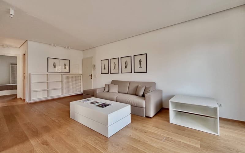 Spacious living room with hardwood floors, white walls, large windows, gray sofa, white coffee table, and framed artwork on the walls