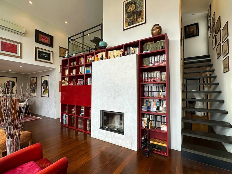Spacious living room with hardwood floors, white walls, and a large built-in bookshelf in a deep red color. The bookshelf is filled with books, decorative items, and a fireplace. The room also features framed artwork on the walls and a red sofa.