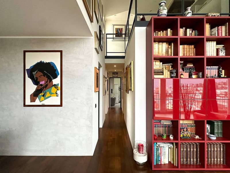 Hallway with white walls, hardwood floors, framed artwork, and a large red bookshelf filled with books and decorative items