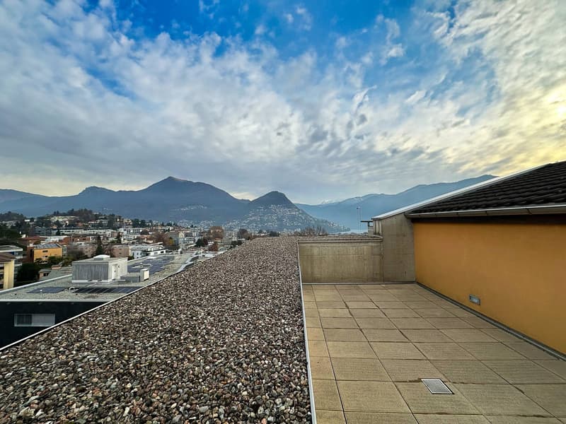 The image shows a rooftop terrace or balcony with a view of a mountainous landscape. The terrace has a tiled floor and is surrounded by a concrete wall. In the distance, there are several buildings and mountains visible under a cloudy sky.