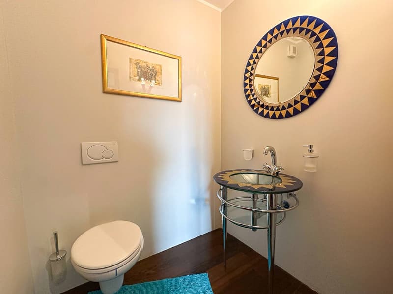 The image shows a bathroom with a modern vanity sink, a round decorative mirror, and a framed artwork on the wall. The bathroom has a toilet and appears to be well-designed with a clean and stylish aesthetic.
