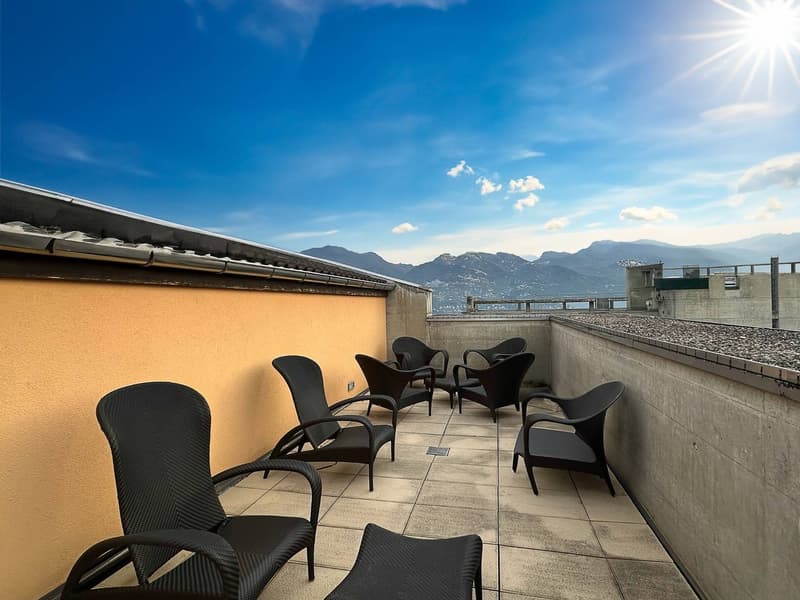 Outdoor balcony or terrace with black wicker furniture, overlooking a scenic mountain landscape with a blue sky and sun shining.