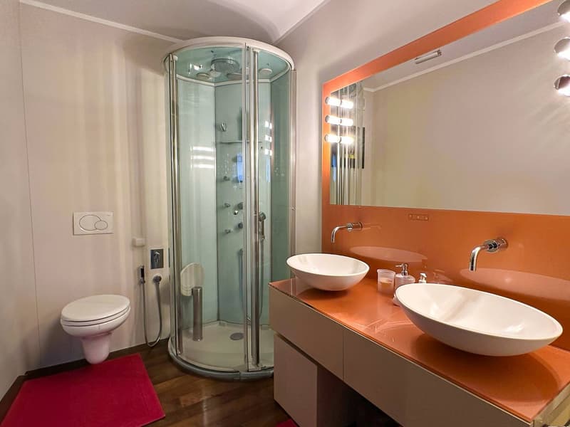 The image shows a modern bathroom with a glass-enclosed shower stall, a vanity with two sinks, and a toilet. The walls are painted in a light color, and the floor appears to be made of hardwood. The bathroom has a sleek and contemporary design with orange