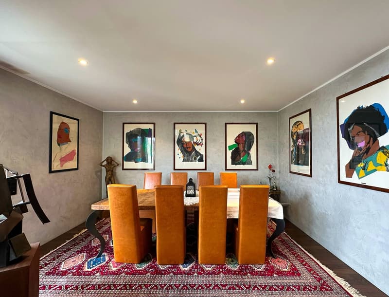 The image shows a dining room with a large wooden dining table surrounded by orange leather chairs. The walls are decorated with colorful abstract art prints in black frames. The room has a warm and modern aesthetic, with a patterned rug on the floor and