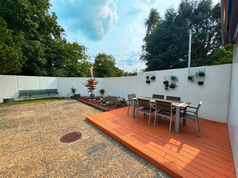 Backyard with wooden deck, outdoor dining table, plants and a white fence, open view to trees