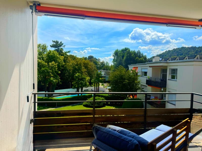 Terrace with metal railings, wooden lounge chairs, cushions, pool view, clear sky with clouds, trees, and mountains in the distance