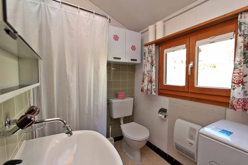 Bathroom with white sink, white shower curtain, white toilet, white cabinets, white washing machine, wooden window, floral curtains, tiled walls, and a toilet paper holder.