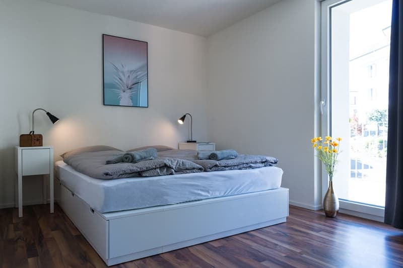 Spacious bedroom with large windows, hardwood flooring, white walls, a queen-size bed with gray bedding, two bedside tables with lamps, and a framed artwork on the wall. There is also a vase with yellow flowers on the windowsill.