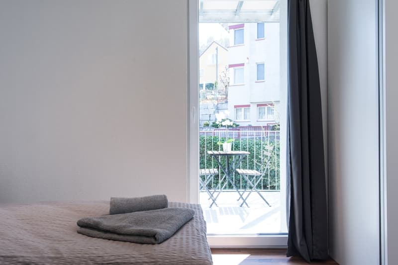 Bedroom with gray bedding, small balcony with chairs visible through the window