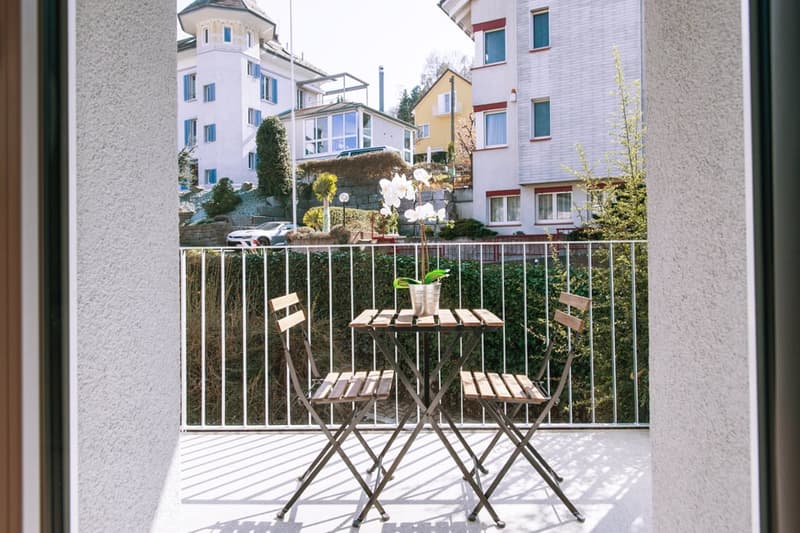 Balcony with a small table and chairs, overlooking a residential area with multi-story buildings and greenery.