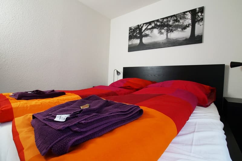 The image shows a bedroom with a black wooden headboard, a large black and white landscape photograph on the wall, and a bed with red and orange bedding. There is also a purple blanket on the bed.