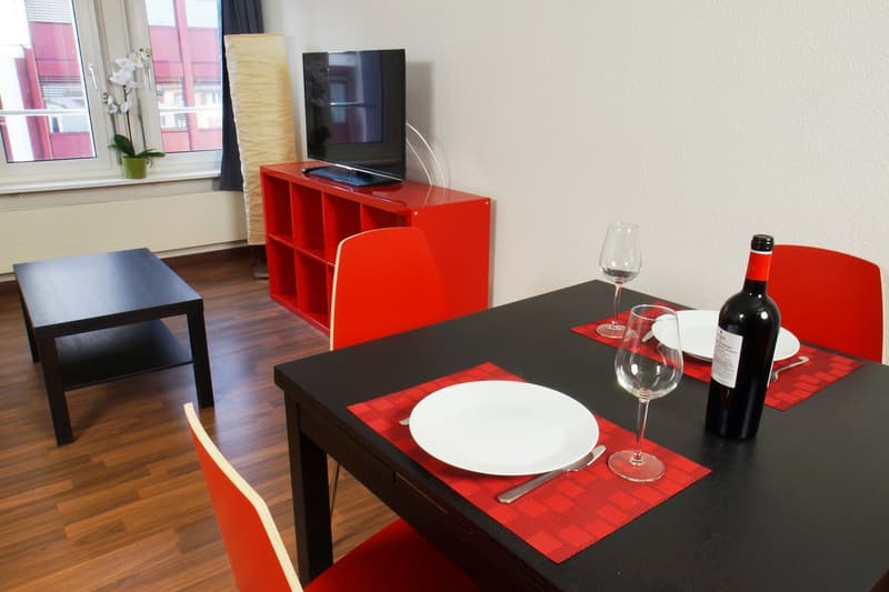 The image shows a modern living room with a red storage unit holding a TV, a black coffee table, and a dining table set with a plate, wine glasses, and a bottle of wine. The room has hardwood floors and large windows.