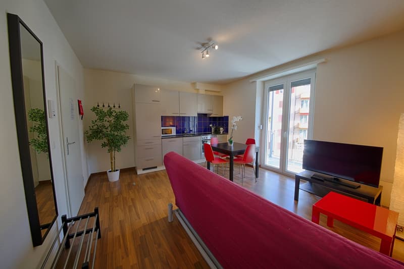 Open-plan living room and kitchen with hardwood floors, white cabinets, blue tile backsplash, dining table with red chairs, large window, and a potted plant. There is also a TV stand and a balcony or terrace visible.