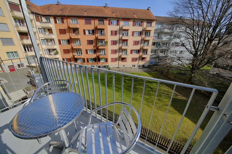 Multi-story apartment building with orange and red brick facade, balconies, and a grassy area in the foreground with a small table and chairs on a balcony.