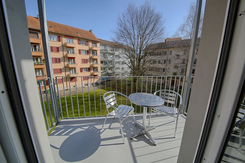 Balcony with a small table and chairs, overlooking a residential area with multi-story apartment buildings and a tree in the foreground.