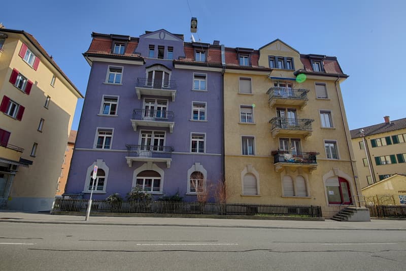 Multi-story apartment building with a mix of yellow and purple facades, featuring balconies and decorative architectural elements.