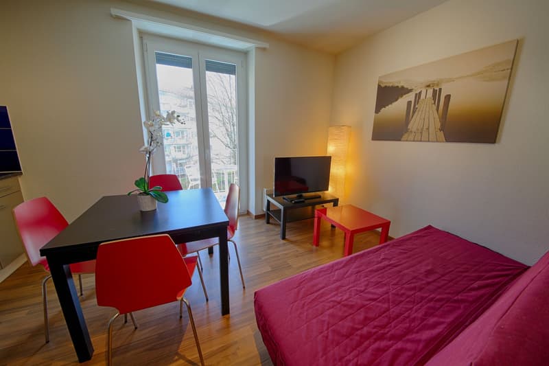 Bright and modern studio apartment with large windows, hardwood floors, dining table with red chairs, TV stand, and a red quilted bed cover.